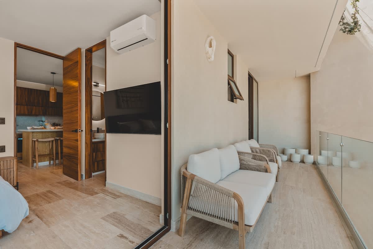 An interior view transitioning to a long balcony with outdoor seating and a stunning ocean view. The room features light flooring, an air conditioning unit, and a glimpse into a modern kitchen with wooden cabinetry through an open doorway.