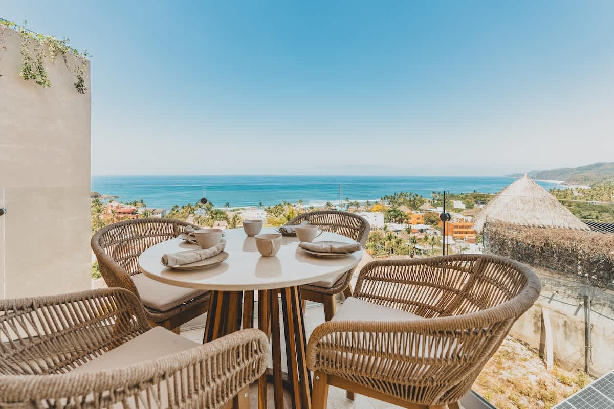 Outdoor dining area on a balcony with a round table set for four, featuring woven chairs and place settings, all overlooking a stunning panoramic ocean and coastal town view.