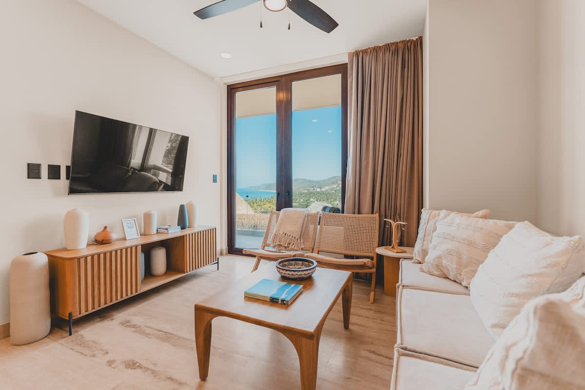 Bright living room with a large wall-mounted TV, a wooden media console, a light-colored sofa, and a wooden coffee table, all facing floor-to-ceiling windows with panoramic ocean and mountain views.