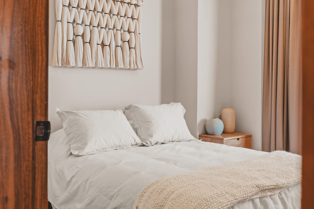 Cozy bedroom featuring a bed with white linens and pillows, a light-colored woven wall hanging above the headboard, and a wooden nightstand with decorative vases.