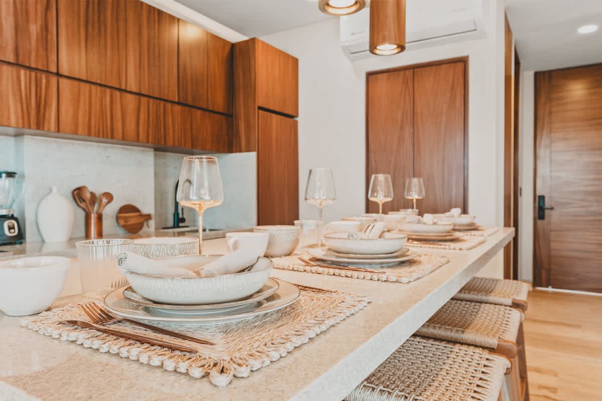 Close-up of a dining counter with a light granite surface, set with four place settings including woven placemats, ceramic dishes, and wine glasses, illuminated by three cylindrical pendant lights.