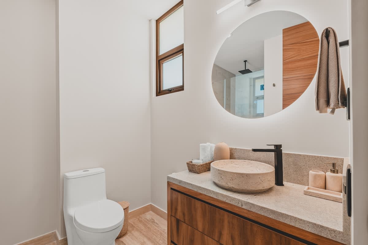 A bright bathroom with a modern toilet and a vanity featuring a light stone countertop, a round vessel sink, and a large circular backlit mirror. A small window provides natural light.