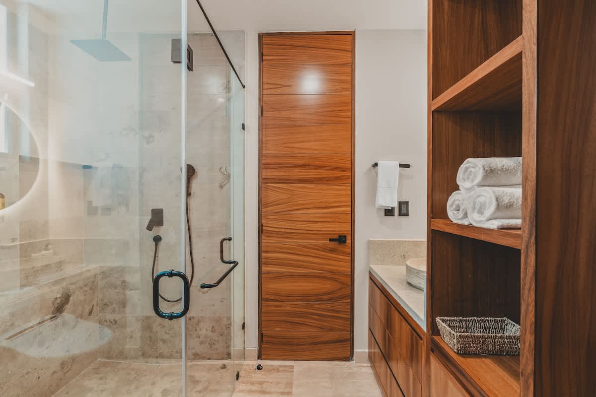 A spacious bathroom featuring a glass-enclosed walk-in shower with stone tiles, adjacent to a modern vanity area with wooden cabinetry and open shelving. A sleek wooden door is also visible.