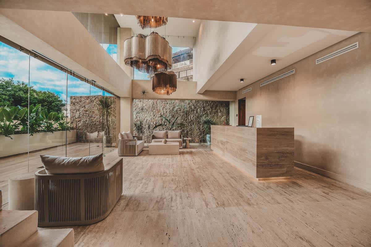 Spacious, modern resort lobby featuring a long, light-toned stone reception desk, a seating area with contemporary sofas and a concrete coffee table, and floor-to-ceiling glass walls overlooking lush tropical foliage. Ornate, multi-tiered chandeliers hang from the high ceiling, illuminating the polished stone floor.