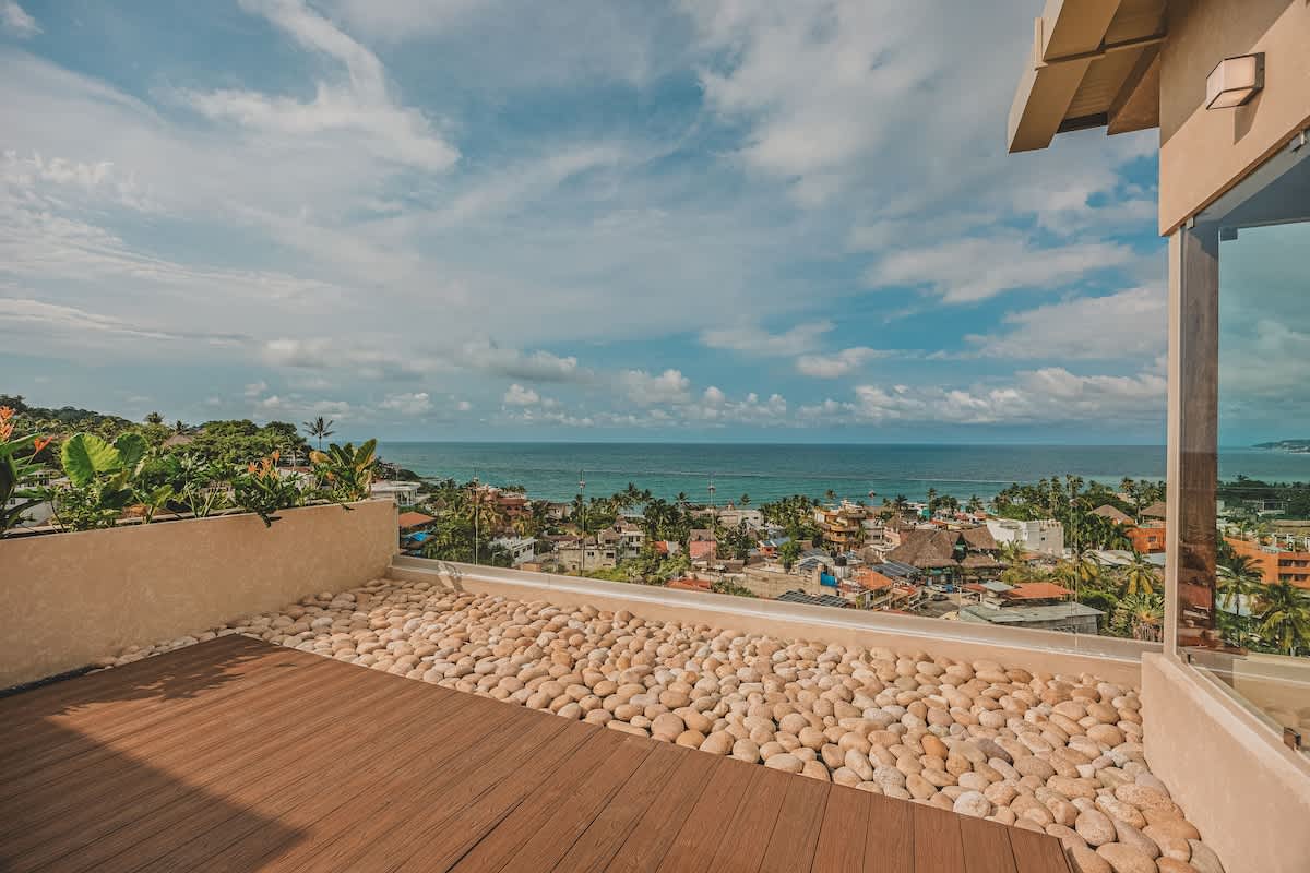 Expansive rooftop deck featuring warm wooden planks and decorative light-colored pebbles, offering a breathtaking panoramic view of a vibrant coastal town, the vast ocean, and lush distant mountains.
