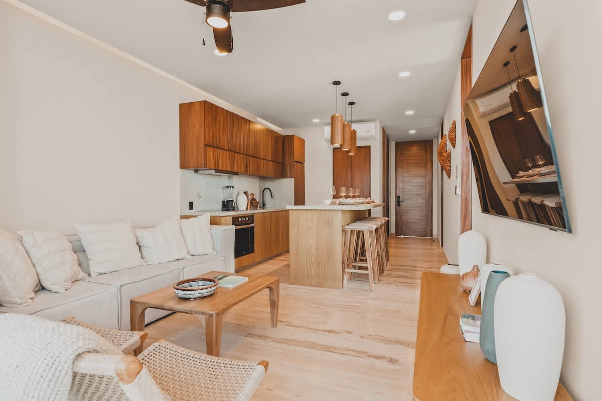 Expansive open-concept living space combining a modern kitchen with a dining island and a comfortable living room, featuring light wood flooring and large windows offering a bright ambiance.