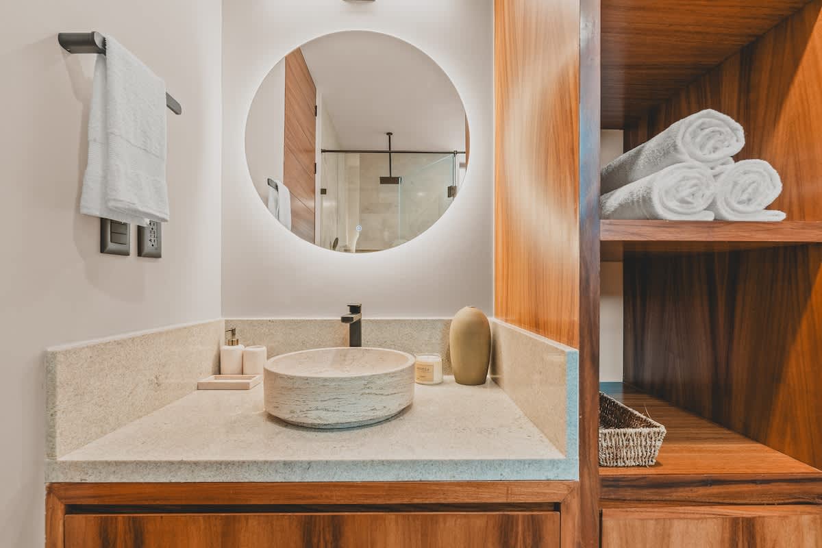 A detailed view of a modern bathroom vanity featuring a light stone countertop, a textured round vessel sink, and a large circular backlit mirror. Black fixtures and neatly folded white towels add to the clean aesthetic.