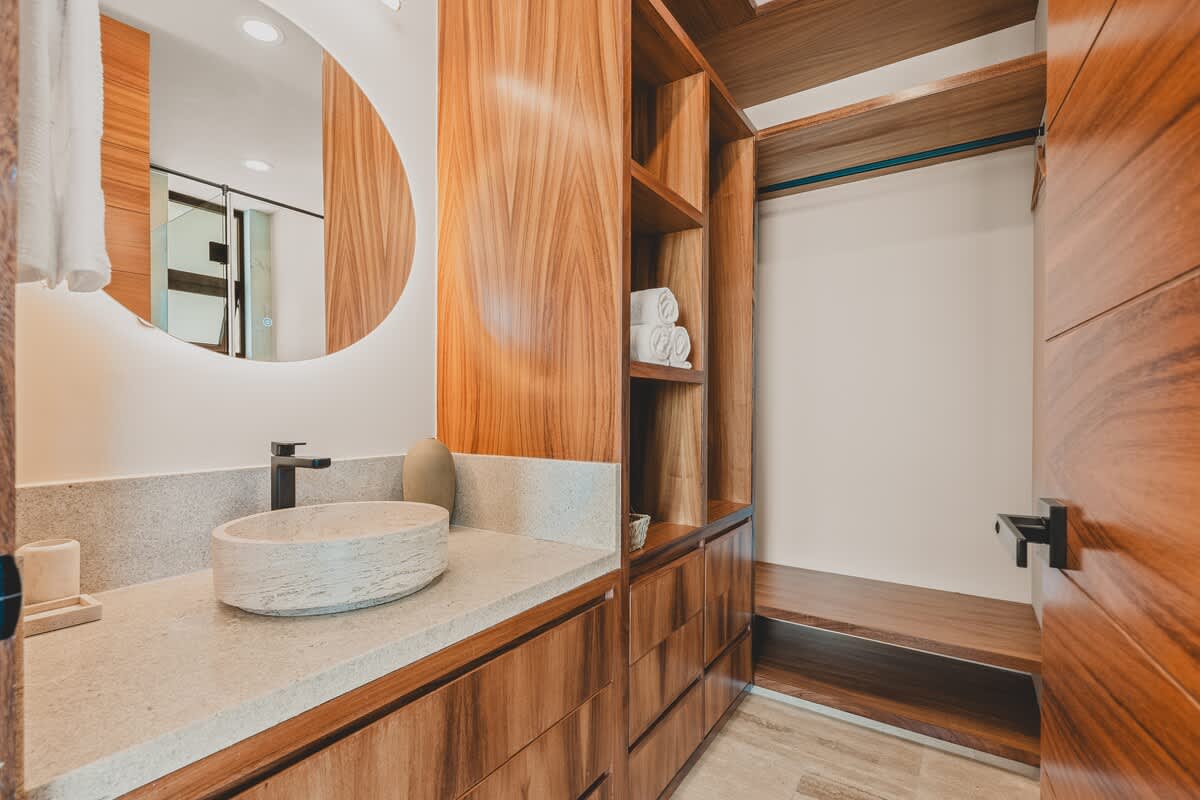A modern bathroom vanity area with a light stone countertop, a round vessel sink, and a large circular backlit mirror. Rich wooden cabinetry and open shelving with rolled towels are prominent features.