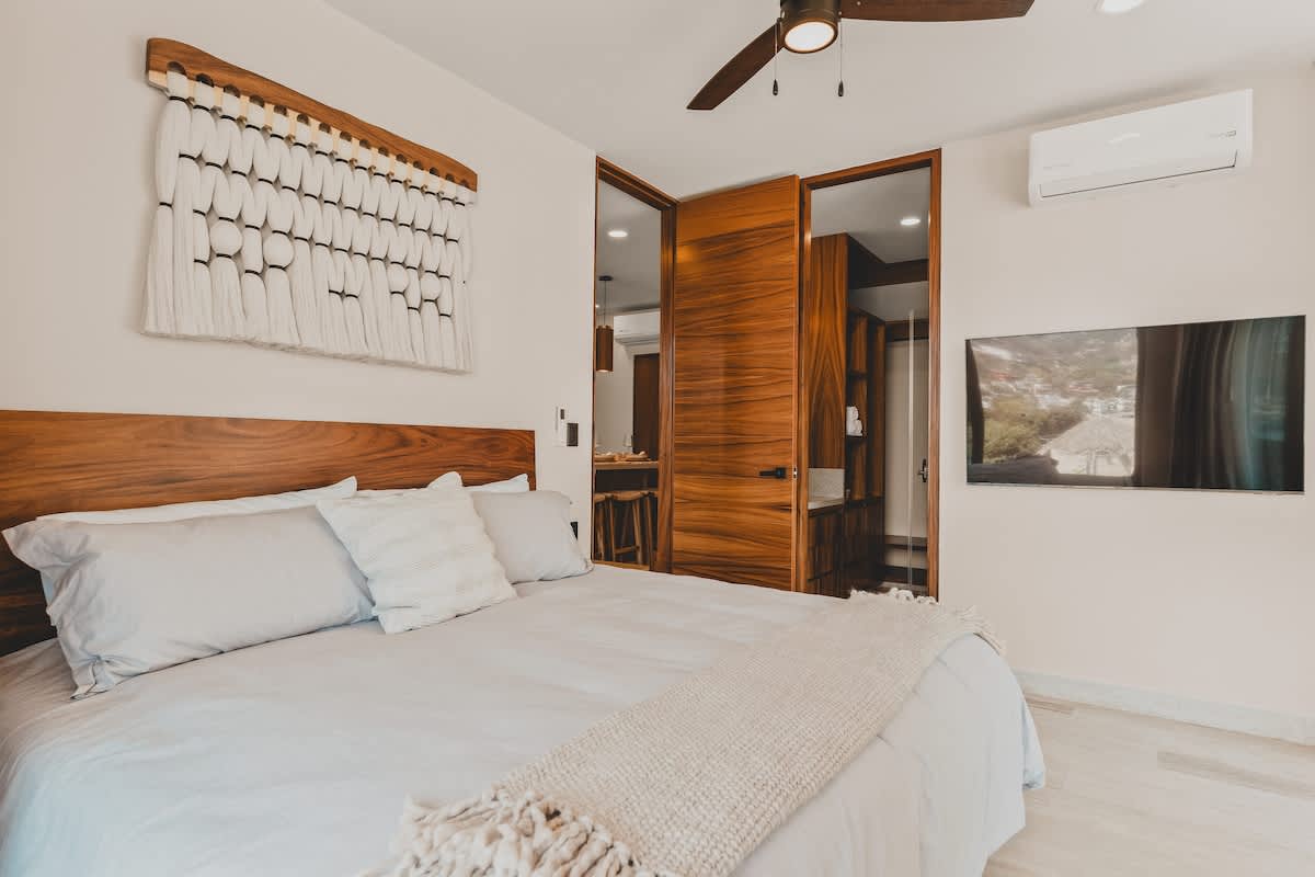 A modern bedroom featuring a large bed with a wooden headboard, light bedding, and a textured white wall hanging above. A ceiling fan and a wall-mounted TV are visible, with a glimpse into a vanity area through an open wooden door.