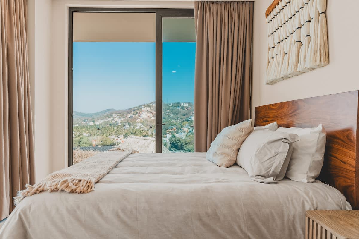 Cozy bedroom with a large bed, a dark wooden headboard, and a sliding glass door providing ample natural light and a scenic view of distant hills and a coastal landscape.