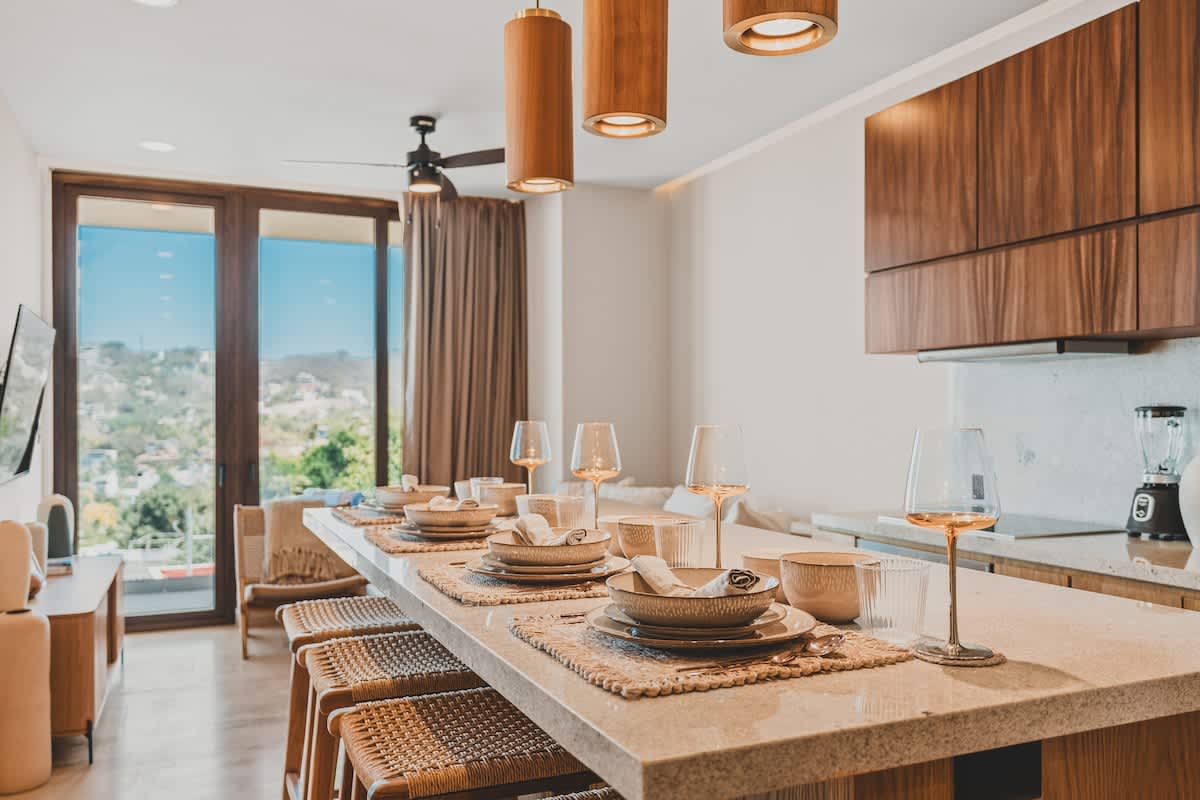 Contemporary kitchen and dining space featuring a long stone island with place settings and woven bar stools, wooden cabinetry, and pendant lighting, with a living area visible in the background.