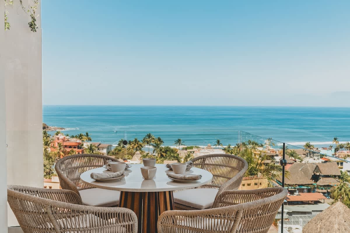 Panoramic view from a balcony featuring a set outdoor dining table with woven chairs, overlooking the expansive blue ocean and the charming rooftops of a coastal town.
