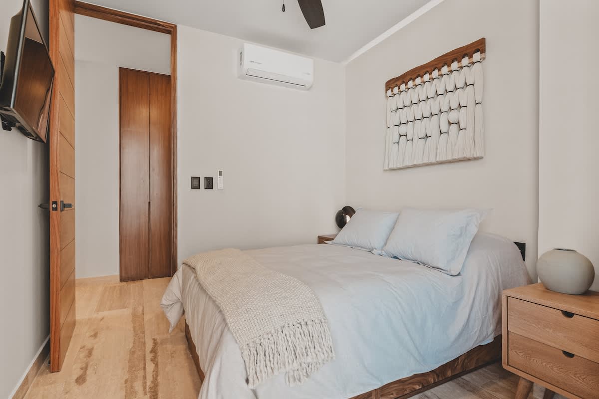 Cozy bedroom with a queen-sized bed, light blue bedding, a wooden headboard, and a textured white wall hanging, complemented by light wood flooring and a minimalist bedside table.