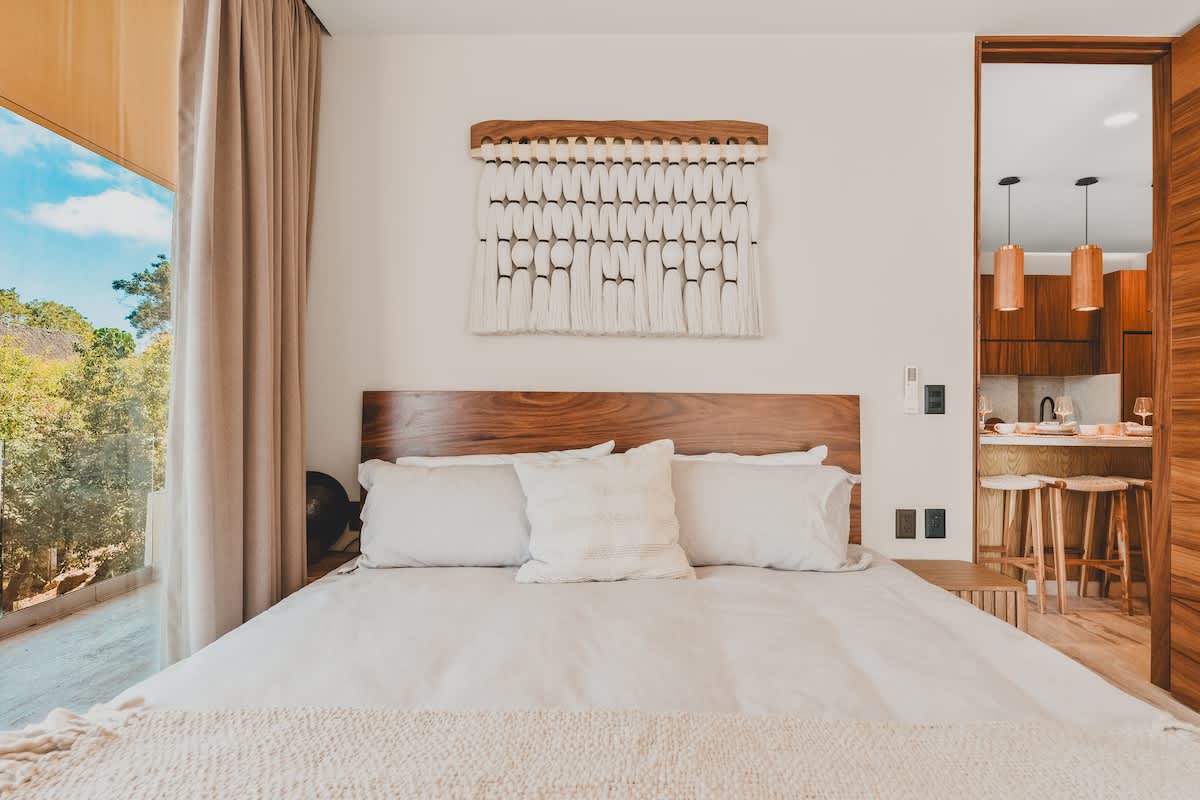 A bright bedroom with a large bed, wooden headboard, and a textured white wall hanging. A large glass door opens to an outdoor view of lush greenery and blue sky, with light-colored curtains.