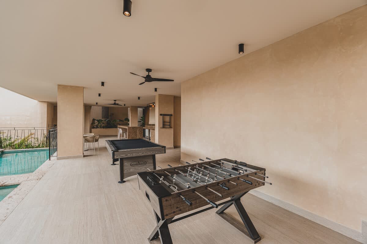 Covered recreation area with light tiled flooring, showcasing a foosball table in the foreground and a pool table further back. The space is open to a swimming pool on the left, with a dark ceiling fan overhead.