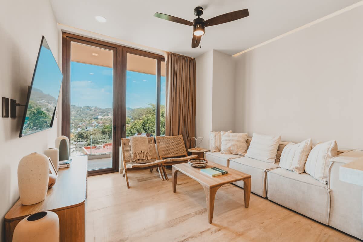 Bright living room with a light-colored sectional sofa, two woven armchairs, a wooden coffee table, and a wall-mounted TV, featuring large glass doors opening to a balcony with a lush green view.