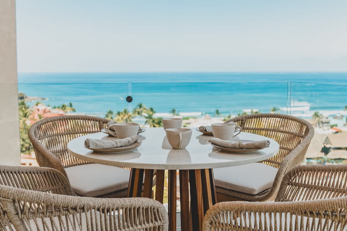Outdoor dining table on a balcony, set with four cups and napkins, offering a stunning, unobstructed view of the vast blue ocean and the horizon.