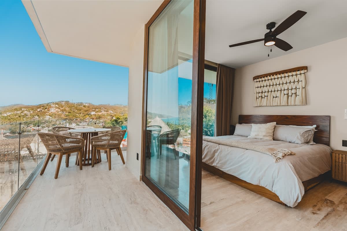 Modern bedroom featuring a wooden bed frame, a ceiling fan, and a large sliding glass door leading to a balcony with a small dining set and scenic views of a coastal town and hills.