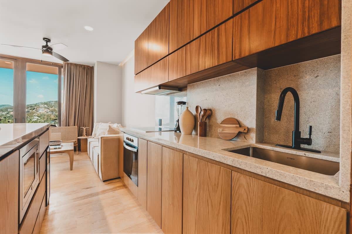 Sleek kitchen with light wood lower cabinets and dark wood upper cabinets, light granite countertops, a black sink, and an induction cooktop, with an open view into the adjacent living area.