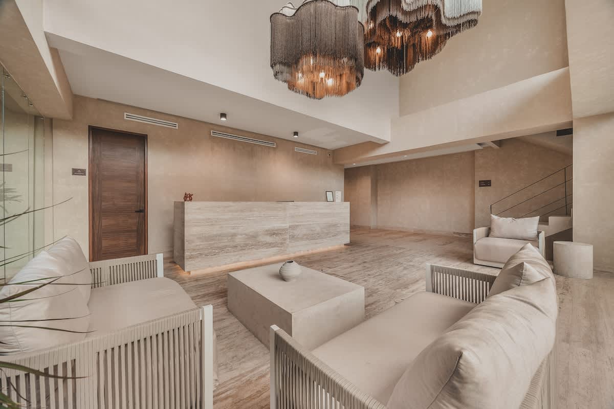 Elegant resort lobby viewed from the lounge area, featuring a long, light-colored reception desk and a modern staircase with dark railings. The double-height ceiling is adorned with striking, tiered chandeliers, casting a warm glow over the polished stone floor.