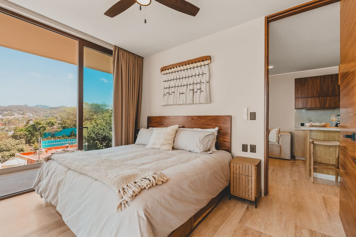 Bright bedroom with a queen-sized bed, light bedding, a wooden headboard, and a textured wall hanging, featuring large glass doors leading to a balcony with a view of lush greenery and distant buildings.
