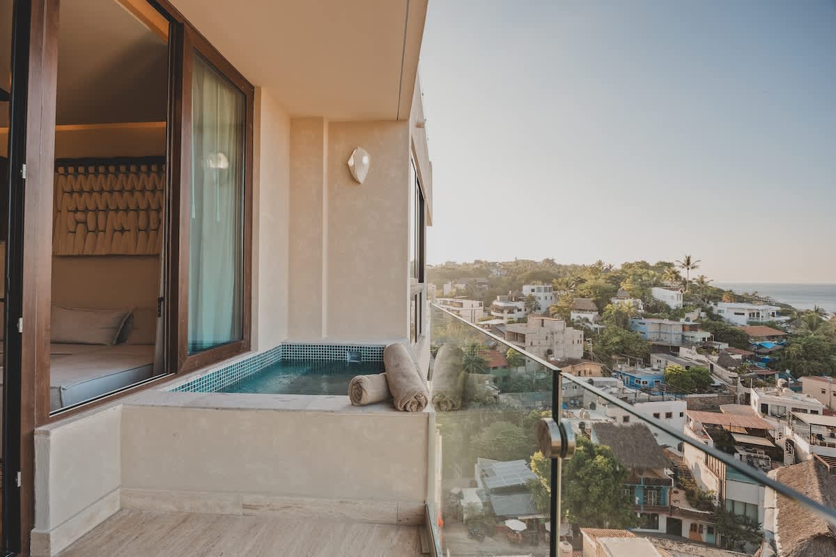 Balcony hot tub with blue mosaic tiles and a glass railing, providing an expansive view of the coastal town and ocean, accessible from an interior room through large sliding glass doors.