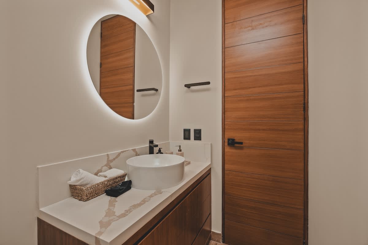 Contemporary bathroom with a dark wood vanity, a white vessel sink, a round backlit mirror, and a tall wooden door, complemented by a white toilet partially visible.