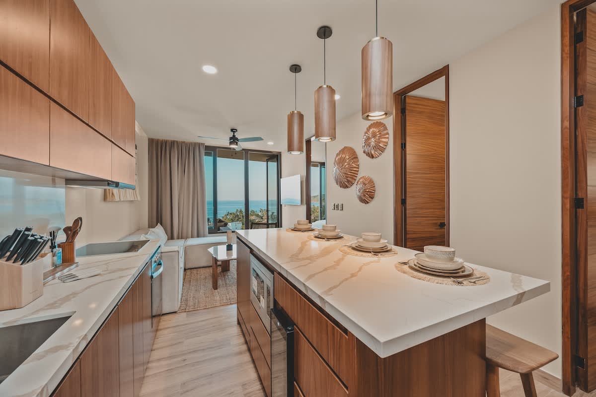 Open-concept kitchen and living area featuring a long kitchen island with place settings, dark wood cabinetry, white marble countertops, and a living space with an ocean view through large glass doors.