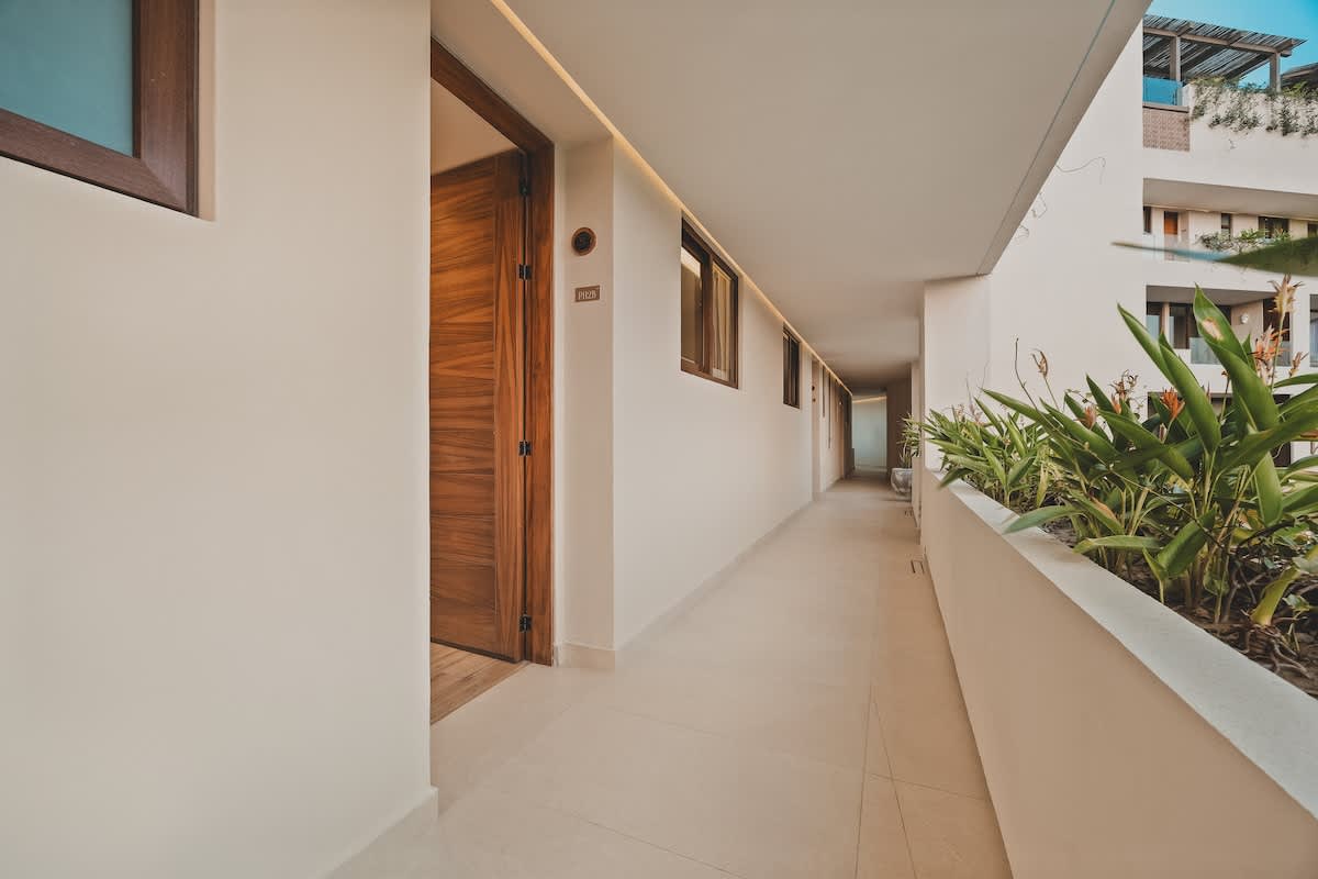 Long, open-air hallway with white walls, light tiled floors, and integrated planters with green foliage, leading past several wooden apartment doors.
