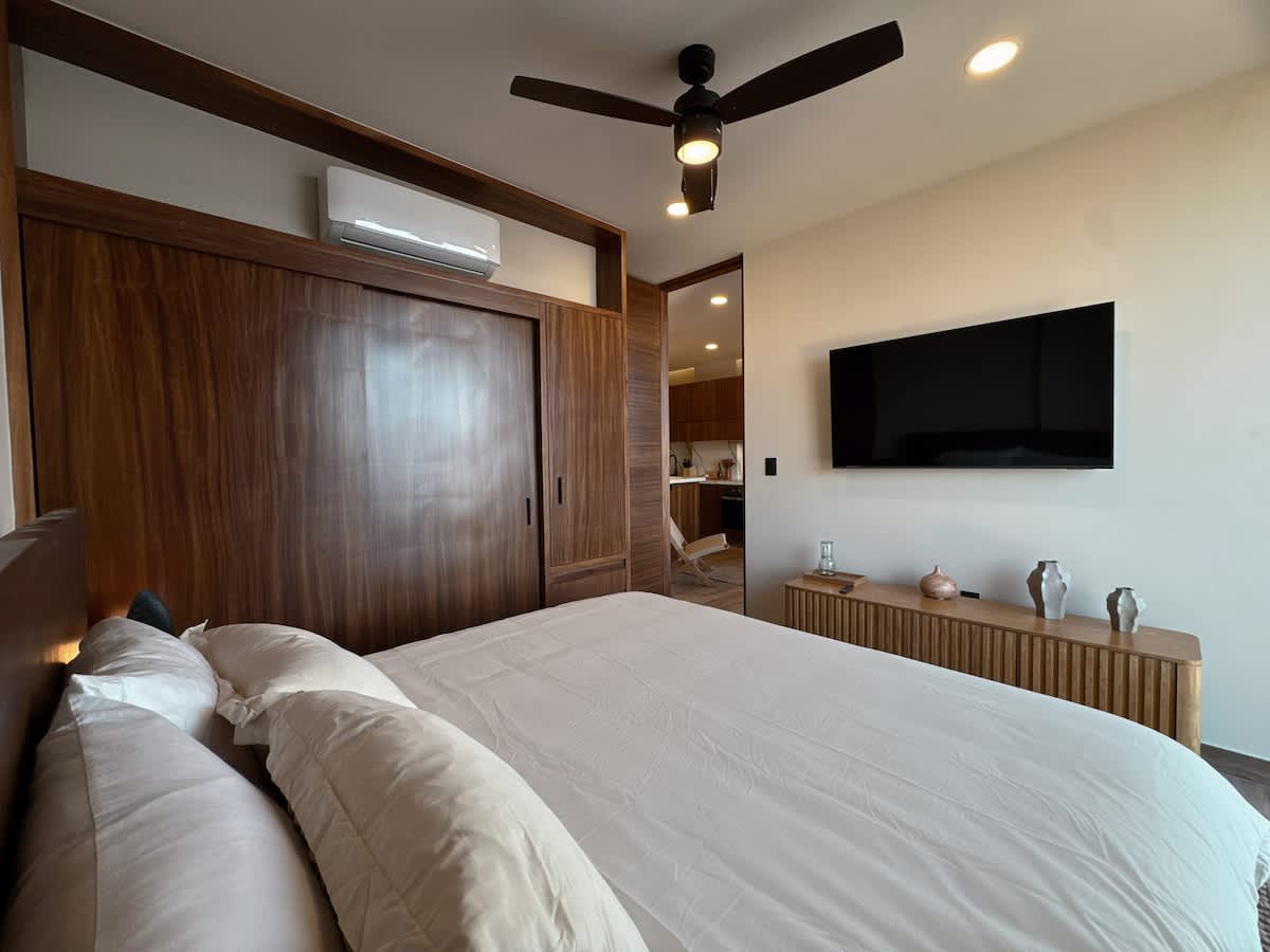 Modern bedroom with a large bed, integrated wooden cabinetry and headboard, a wall-mounted flat-screen TV, and an air conditioning unit, featuring light-colored bedding and decorative vases on a console.