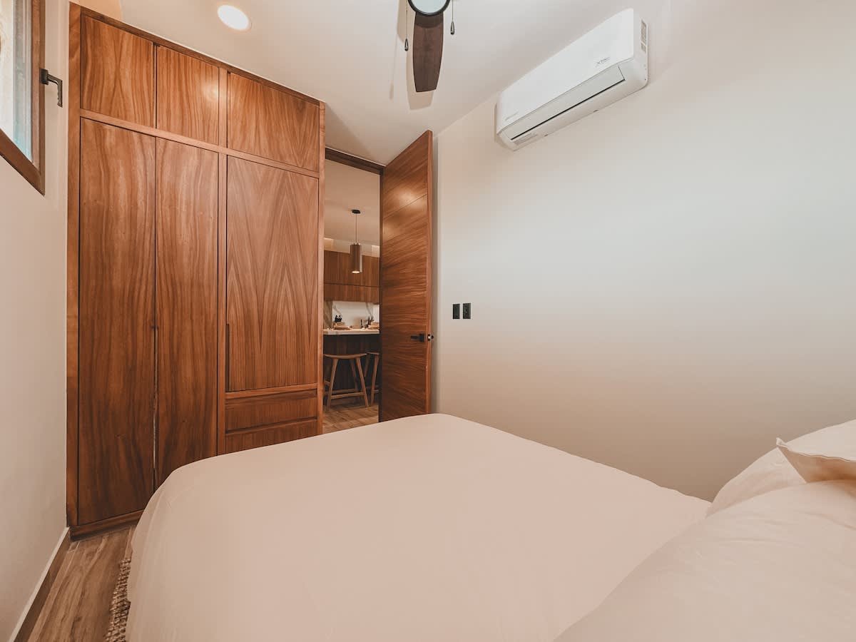 Cozy bedroom featuring a light-colored bed with a simple headboard, a large built-in wooden wardrobe, and a ceiling fan, with a partial view into an adjacent kitchen area through an open doorway.