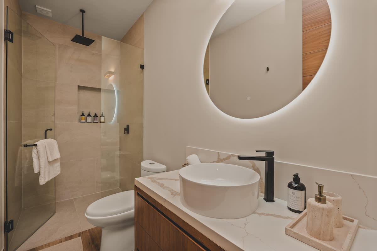Contemporary bathroom vanity with a white vessel sink, black faucet, and a large round backlit mirror, set against a light-colored wall with a wooden vanity cabinet.