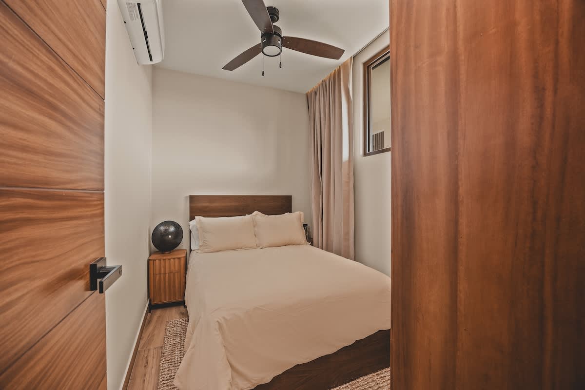 Compact bedroom with a light-colored bed, a wooden headboard, a small wooden nightstand with a decorative sphere, and a built-in wooden wardrobe, featuring a ceiling fan and air conditioning unit.