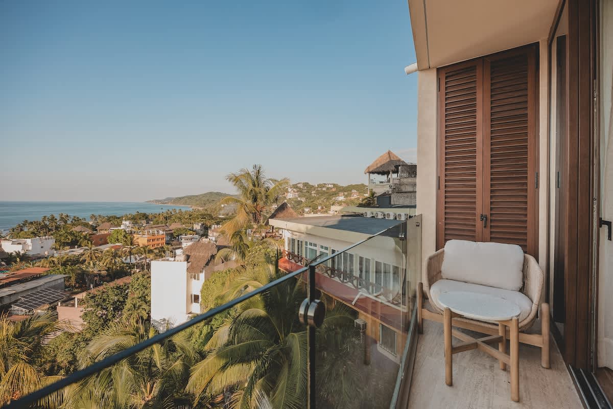 Private balcony with a comfortable woven armchair and a small table, offering a picturesque view of the town's rooftops, palm trees, and the distant ocean. Wooden shutters add a touch of privacy and style.