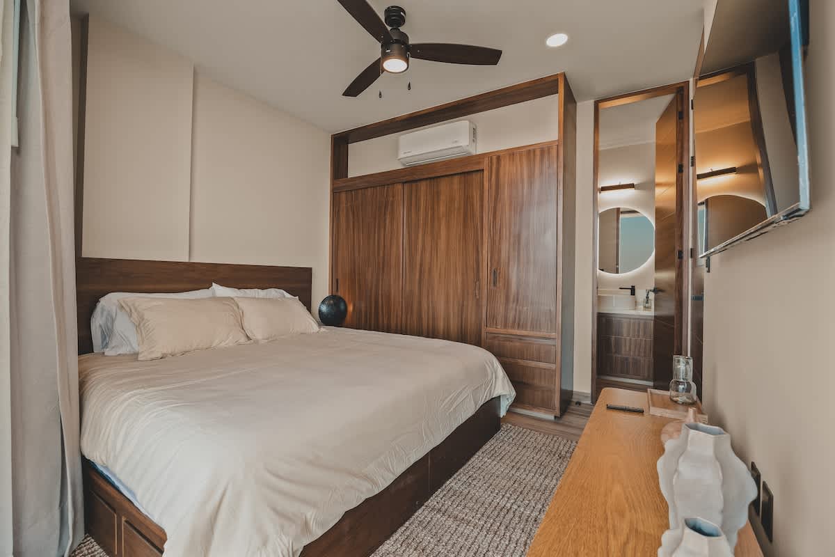 Stylish bedroom featuring a large bed with a wooden headboard, integrated wooden wardrobe and shelving, a wall-mounted TV, and a ceiling fan, with a view into a modern en-suite bathroom.