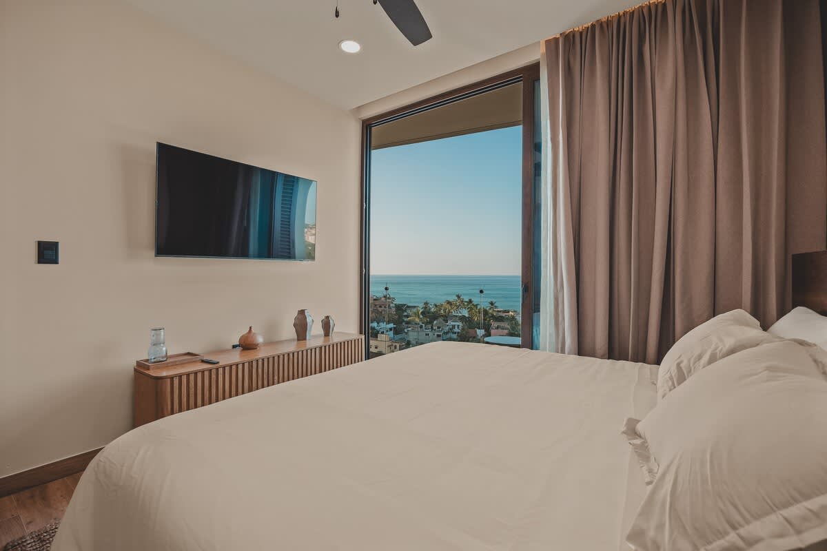 Master bedroom with a large bed dressed in white linens, facing a wall-mounted flat-screen TV above a wooden console. Expansive sliding glass doors offer breathtaking ocean and town views.
