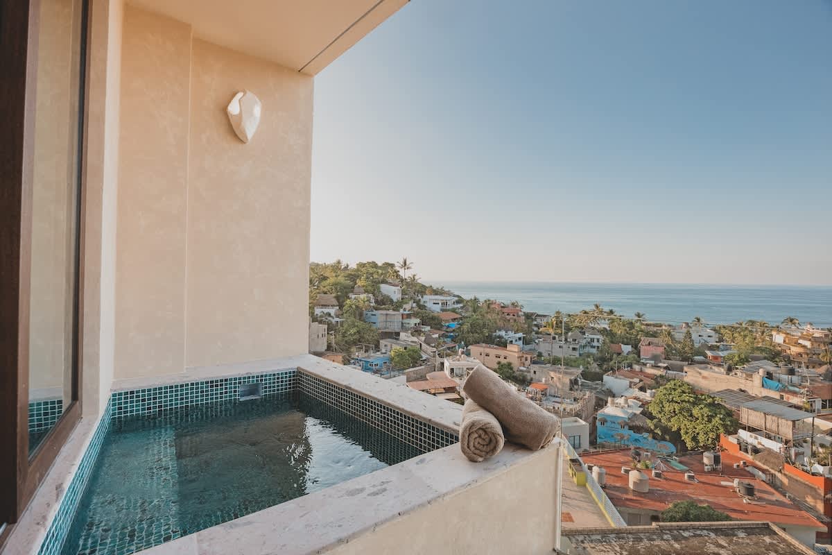 Private balcony hot tub with blue mosaic tiles and a concrete surround, offering stunning panoramic views of a vibrant coastal town with red-tiled roofs and the vast blue ocean under a clear sky.
