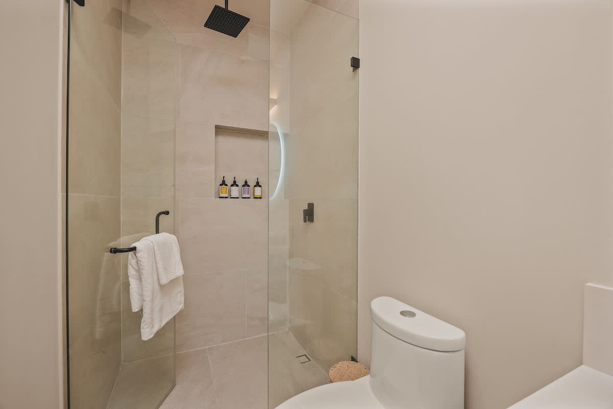 Modern walk-in shower with light beige tiles, a glass enclosure, and a black rain showerhead, adjacent to a white toilet and a vanity with a round backlit mirror.