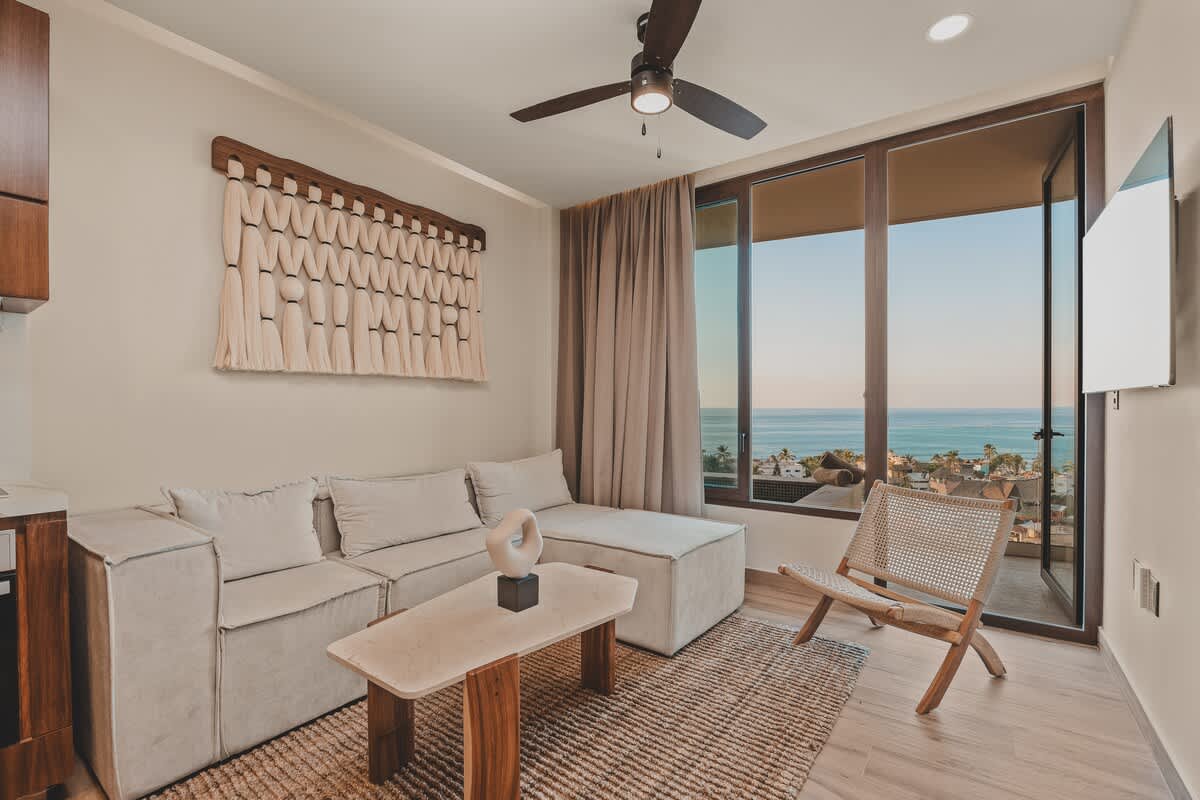 Inviting living room with a light-toned sectional sofa, a unique marble and wood coffee table, and a textured wall hanging. Large glass doors provide abundant natural light and stunning ocean views.