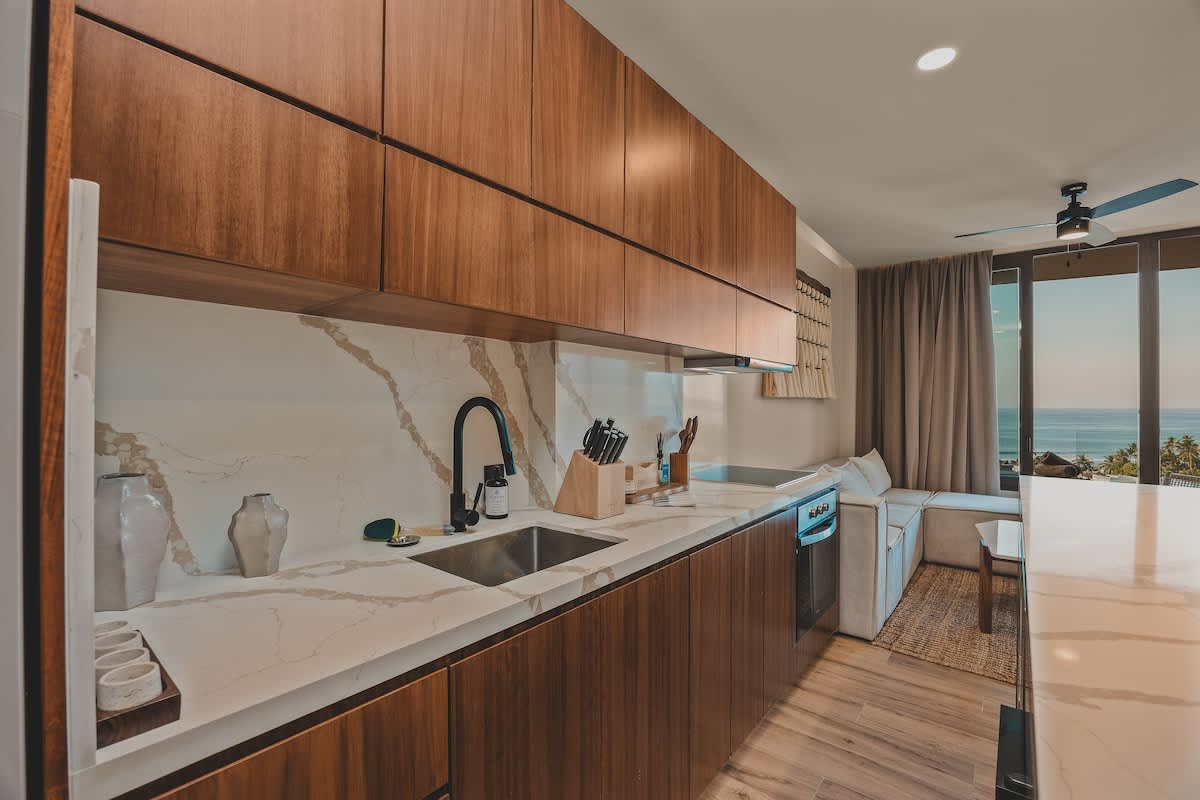 Sleek kitchen with dark wood lower and upper cabinets, white marble countertops and backsplash, a stainless steel sink with a black faucet, and a glimpse of the living area with an ocean view.