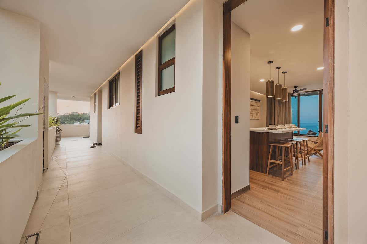Bright hallway with light-colored tiled floors and white walls, leading into an open-concept living space with wood flooring and a kitchen island, offering a glimpse of the ocean.