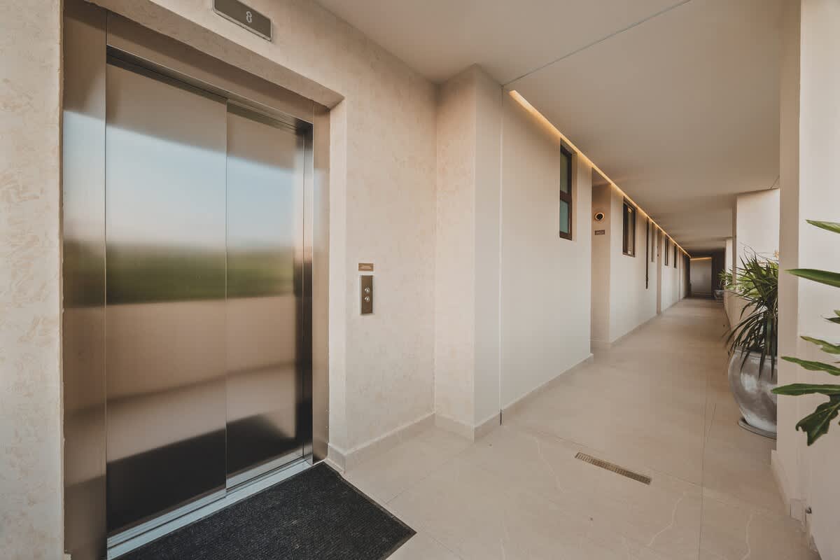 Modern hallway with light-colored walls and tiled floors, featuring a sleek silver elevator with a black mat in front, and a long corridor extending into the distance.