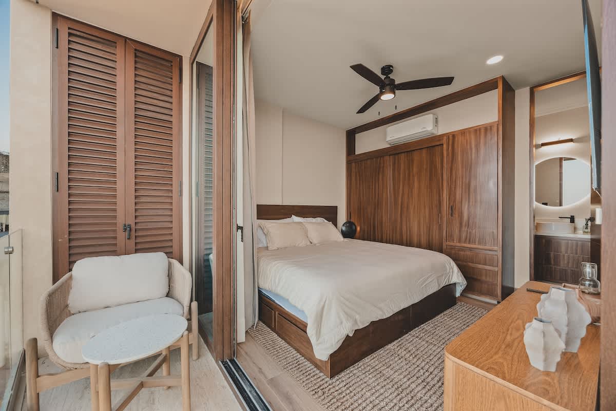 Bright bedroom with a comfortable bed, extensive wooden built-in storage, and direct access to a small balcony with a woven chair and table, featuring a ceiling fan and light-colored curtains.