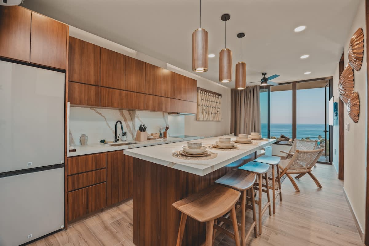 Bright open-plan kitchen and living space with dark wood cabinets, a white marble island, and a large refrigerator, leading to a living area with an ocean view and a ceiling fan.