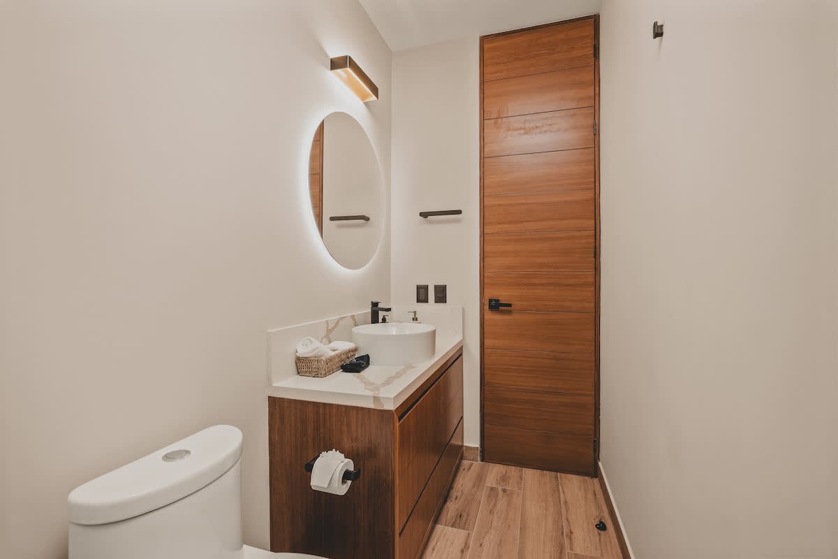 Modern bathroom featuring a white toilet, a dark wood vanity with a white countertop, a round backlit mirror, and a tall wooden door, all set against light-colored walls.