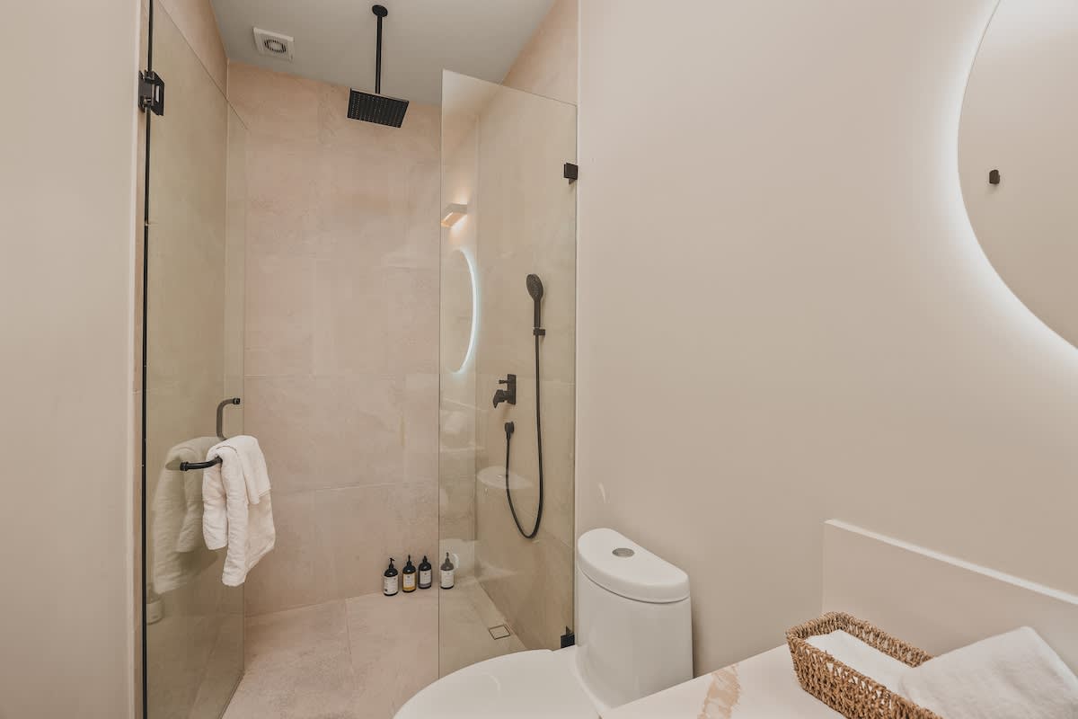 Modern bathroom featuring light beige tiled walls and floor, a glass-enclosed shower with a black rain showerhead, and a white toilet. Small bottles of toiletries are neatly arranged in a shower niche.