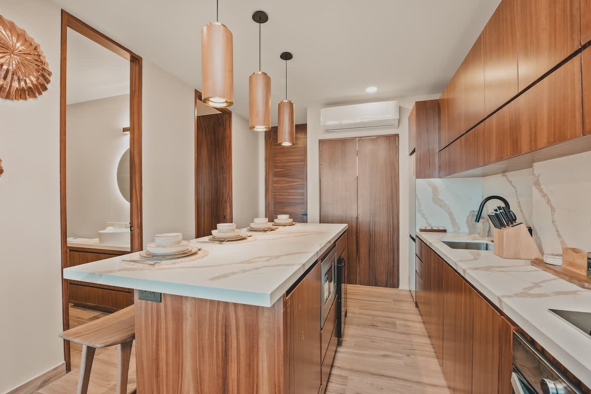 View of the kitchen island with pendant lights and wooden bar stools, showing the entrance door and a wall-mounted air conditioning unit above the door.