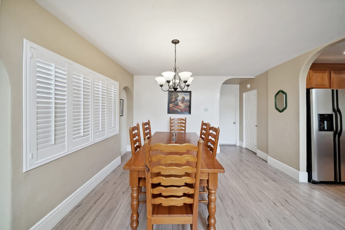 A spacious dining room features a large wooden table with eight matching chairs, illuminated by a chandelier, with light wood flooring and windows covered by white plantation shutters.