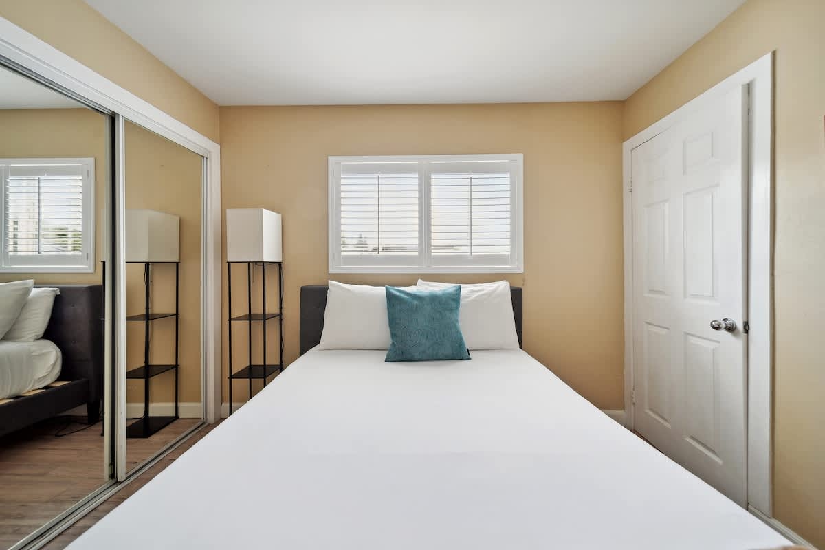 A queen bedroom with light wood flooring features white bedding, a teal accent pillow, a window with white plantation shutters, and a tall black shelving unit.