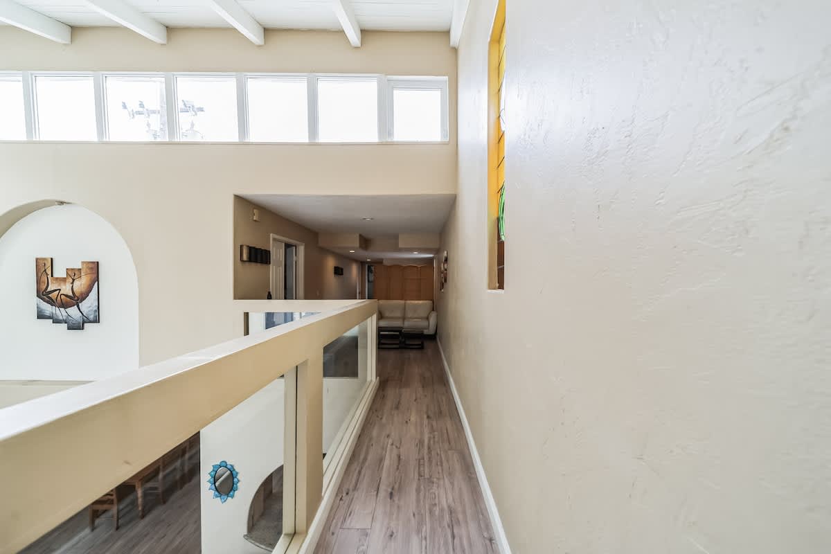 Upper-level hallway or landing with light wood-look flooring, a modern railing with glass panels overlooking a lower floor, and a long corridor extending into the distance.
